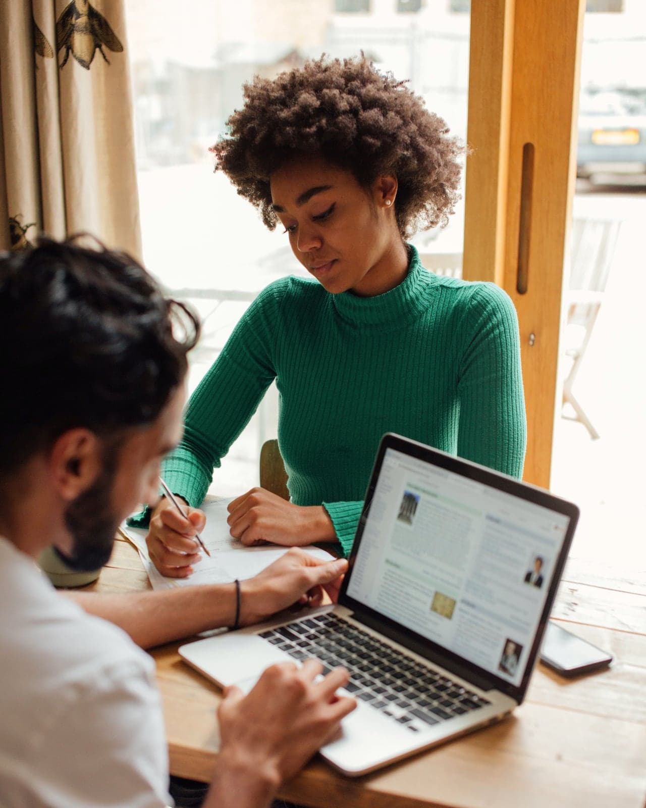 Dos personas trabajando juntas en una mesa junto a la ventana: una toma notas y la otra usa una computadora portátil.