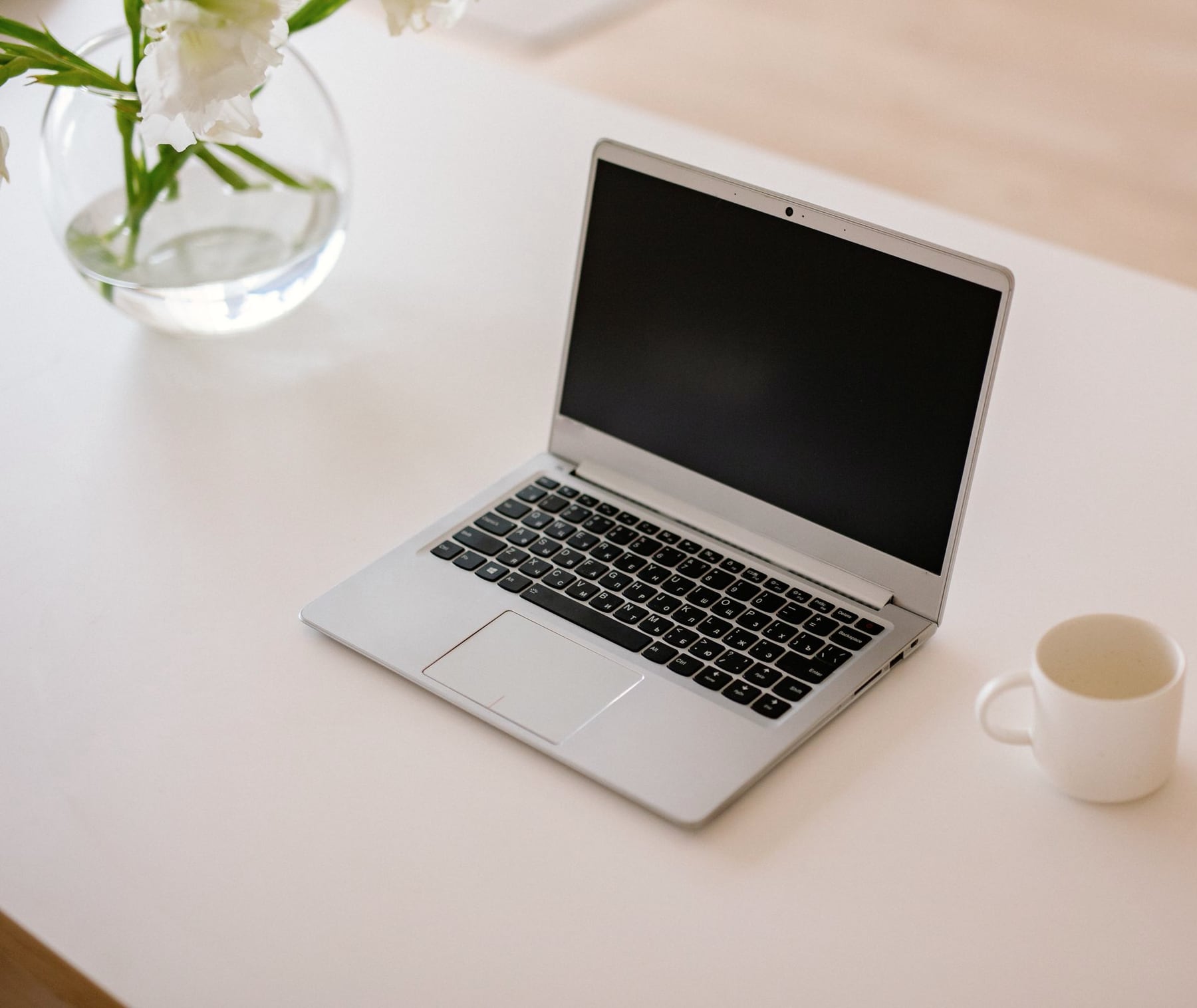 Computadora portátil abierta sobre una mesa clara, vista en tres cuartos, junto a una taza y un florero.